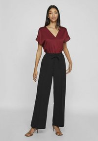 Blouse bordeaux en tissu doux à manches courtes avec un décolleté en V, associée à un pantalon noir taille haute à jambes larges et des sandales à talons bruns à lanières.