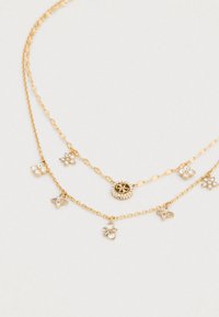 Κολιέ με χρυσά στρώματα που διαθέτει διακοσμητικά μενταγιόν, συμπεριλαμβανομένων λουλουδιών και ενός κυκλικού λογότυπου με διακοσμητικά από στρας.