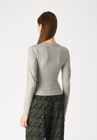 Woman with long brown hair wears fitted light gray long-sleeve top and dark patterned wide-leg pants, standing against white background.