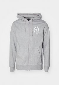 Un sweat à capuche zippé gris avec une poche avant, une capuche à cordon et un logo blanc sur la poitrine gauche. Fabriqué en tissu doux et texturé.