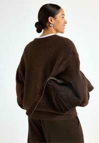 Maglione marrone, oversize, in morbido knit, abbinato a una borsa in suede marrone scuro con una lunga tracolla e un design semplice.