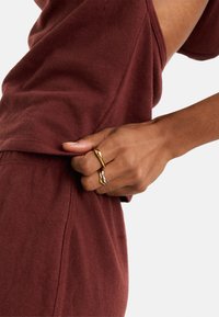 Bordowa, bawełniana bluzka o luźnym kroju z widocznym szwem; ręka trzyma złotą i srebrną, skręconą metalową pierścionek.