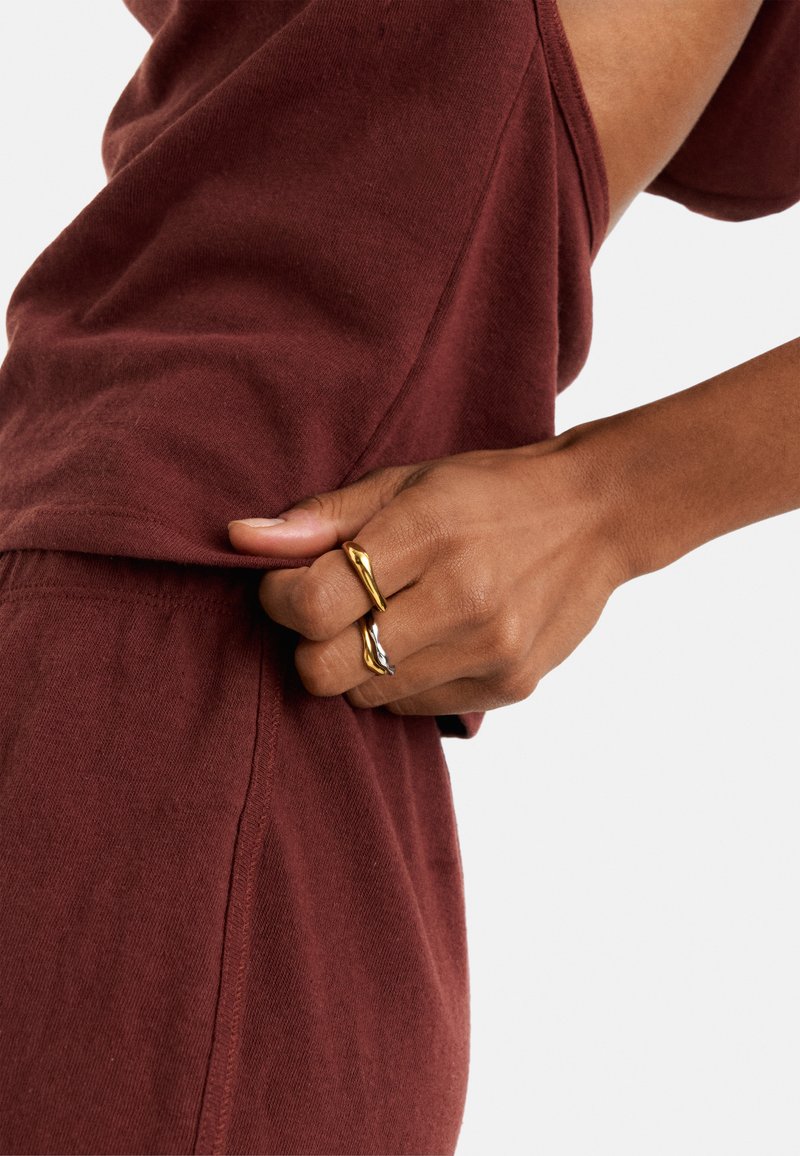 Bordowa, bawełniana bluzka o luźnym kroju z widocznym szwem; ręka trzyma złotą i srebrną, skręconą metalową pierścionek.