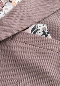 Strukturierter brauner Anzugstoff mit einem gemusterten floralen Einstecktuch, das aus einer Brusttasche hervorblitzt und helle sowie dunkle Farben aufweist.
