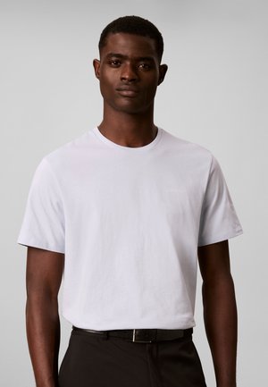 Mann trägt ein schlichtes weißes T-Shirt und schwarze Hose mit Gürtel, steht vor einem hellgrauen Hintergrund.