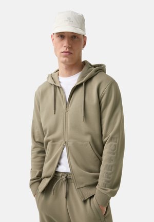 Olive green zip-up hoodie with kangaroo pockets, ribbed cuffs, and logo detail on the sleeve. Worn over a white t-shirt and matching pants.