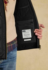 Wnętrze czarnej pikowanej kurtki z zasuwaną kieszenią i metką z instrukcją pielęgnacji. Kurtka ma złote akcenty zamka błyskawicznego oraz teksturowane wykończenie.