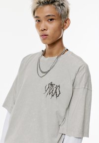 Maglietta oversized grigia chiara con un design grafico nero invecchiato sul petto, tessuto testurizzato e collo a taglio grezzo. Collana in argento.