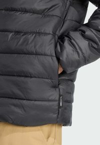 A close-up of a person wearing a dark puffer jacket with visible quilted sections, hands inside pockets, and a label reading “climawarm.”