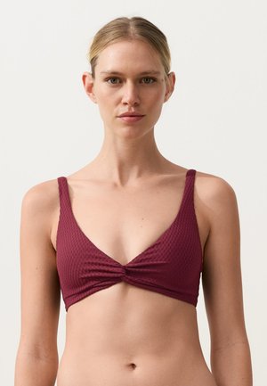 Ženska, ki nosi teksturiran, temno rdeč bikini zgornji del z ozkimi naramnicami in zvitim sprednjim detajlom, stoji pred enobarvnim belim ozadjem.