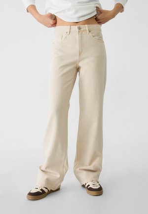 Osoba nosząca kremowe jeansy o szerokich nogawkach i brązowo-białe sneakersy, trzymająca się obiema rękami za talię na jednolitym tle.