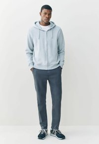 Szürke cipzáras kapucnis pulóver, húzózsinóros kapucnival, fehér ing fölött viselve, sötétszürke nadrággal és fekete sportcipővel párosítva.