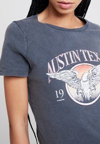Camiseta de algodón gris con un estampado gráfico redondo que presenta un águila y el texto "AUSTIN TEX" en colores desvaídos. Manga corta y escote redondo.