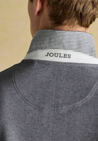 Šedá polo košeľa s pruhovaným golierom. Logo "JOULES" v tmavej farbe na bielom podklade. Hladká textúra s jemnými švovými detailmi.