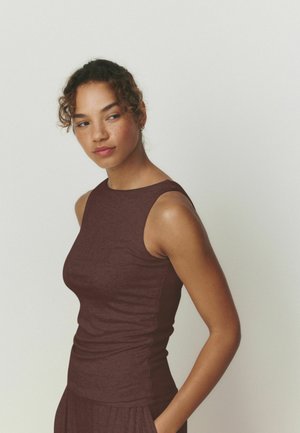 Junge Frau mit lockigem Haar in einem ärmellosen bordeauxfarbenen Oberteil und Rock, seitlich stehend mit den Händen in den Taschen, schaut nach links.