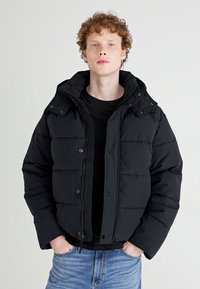 Chaqueta de plumas negra con capucha, que cuenta con un cierre de cremallera y botones a presión, paneles laterales acanalados y textura acolchada. Lleva puesta una camiseta negra.