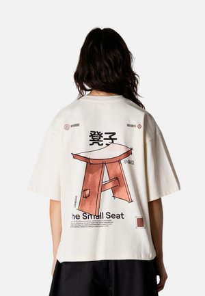 Person med langt mørkt hår iført en hvid oversized T-shirt med en stor illustration af en brun træskammel og japansk tekst på bagsiden.