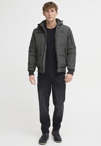 Polstret sort jakke med hætte, lynlås foran og ribbede manchetter. Bæres over en sort skjorte og parres med mørke jeans og støvler.