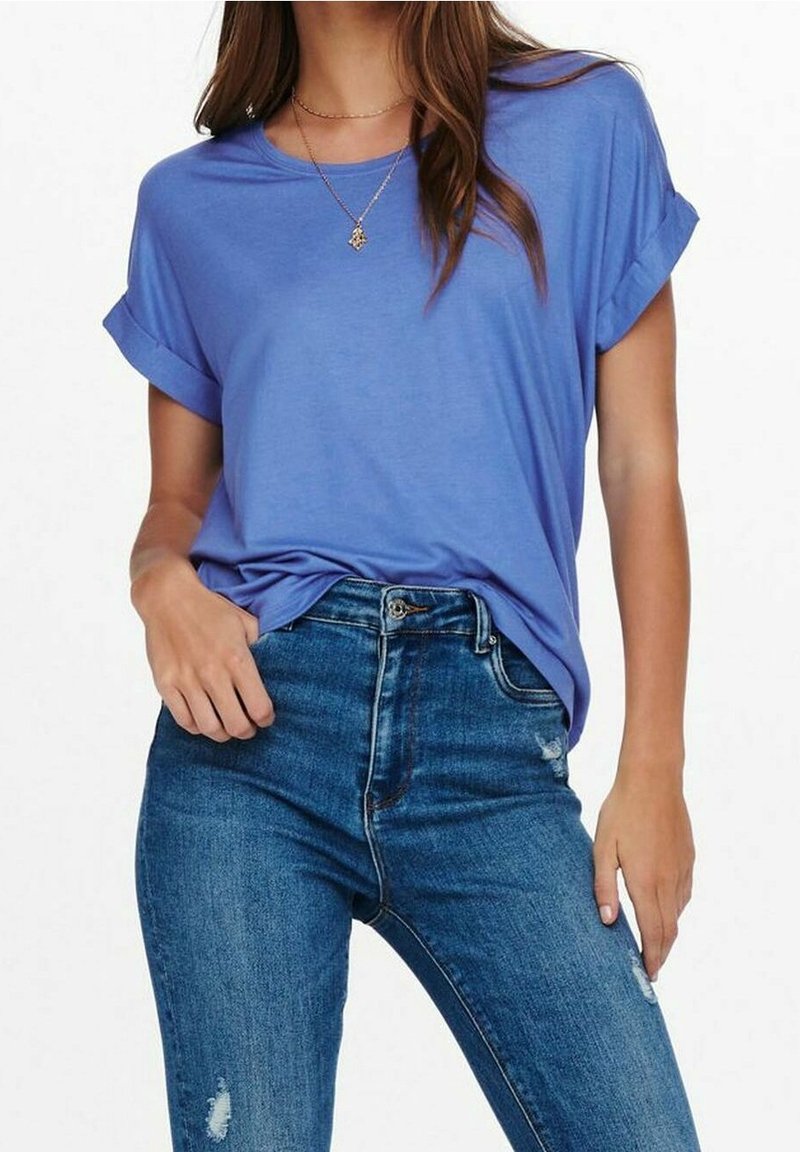 Femme portant une chemise bleue ample à manches courtes légèrement rentrée dans un jean bleu taille haute avec une légère usure, debout devant un fond uni.