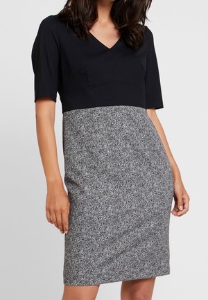 Femme portant une robe longueur genou avec le haut noir et une jupe grise à motif chevrons, debout devant un fond uni.