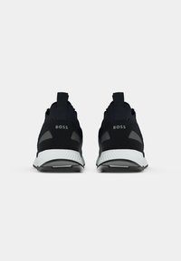 Paire de chaussures de sport noires avec semelles blanches, vues de dos, chacune marquée du mot BOSS sur la languette du talon.