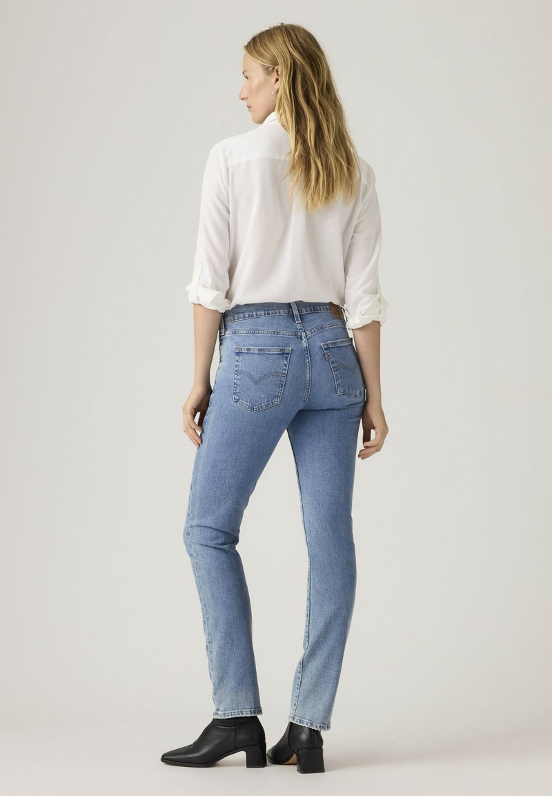 Kvinde set bagfra iført lyseblå højtaljede jeans, hvid langærmet skjorte med opsmøgede ærmer og sorte ankelstøvler på neutral baggrund.