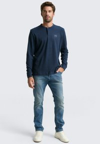 Mann steht mit der Hand in der Tasche, trägt ein navyfarbenes Langarmhemd, hellblaue Jeans und weiße Sneakers vor einem schlichten Hintergrund.