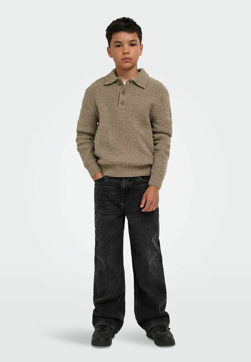 Garçon debout, une main dans la poche, portant un pull en laine beige, un jean noir ample et des chaussures noires sur un fond clair uni.