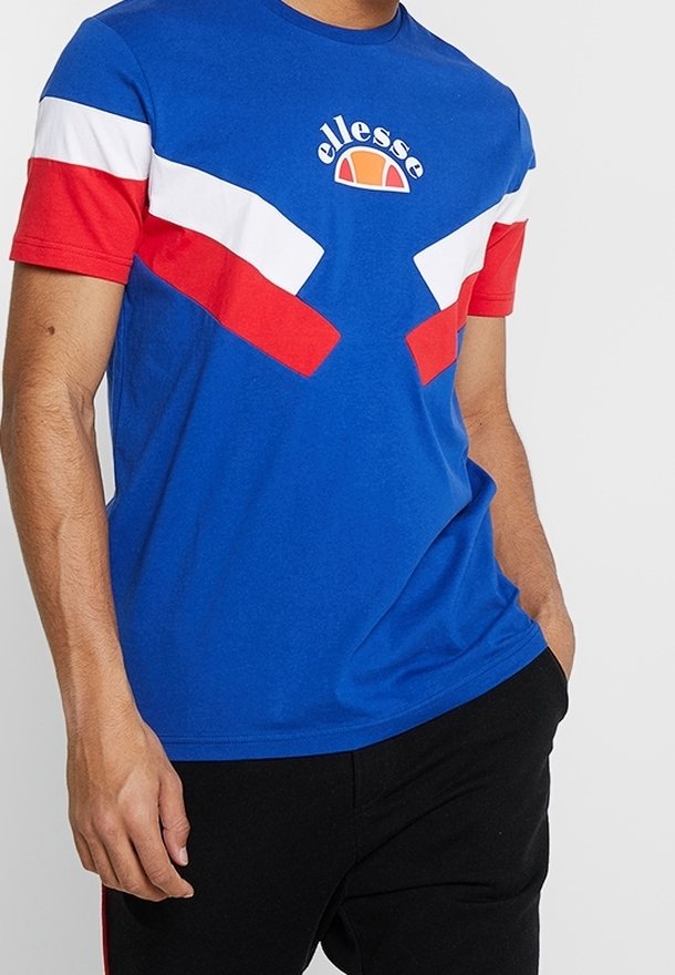 Homme portant un T-shirt à manches courtes bleu, rouge et blanc avec un motif géométrique et le logo « ellesse » sur la poitrine, les mains dans les poches.
