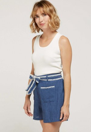 Femme aux cheveux blonds portant un haut blanc sans manches et un short bleu avec une ceinture nouée assortie et une bordure décorative.