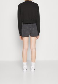 Černá zkrácená mikina, tmavě modré džíny s vysokým pasem a roztrhaným lemem, bílé ponožky Nike a tenisky, jednobarevné pozadí.