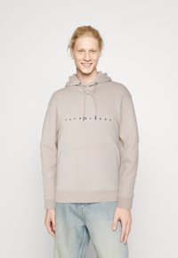 Šviesiai smėlinė vilnonių striukė su raišteliu, turinti priekinę kengūros kišenę ir juodą „JACK & JONES“ logotipą, įsiterpusį į krūtinę.