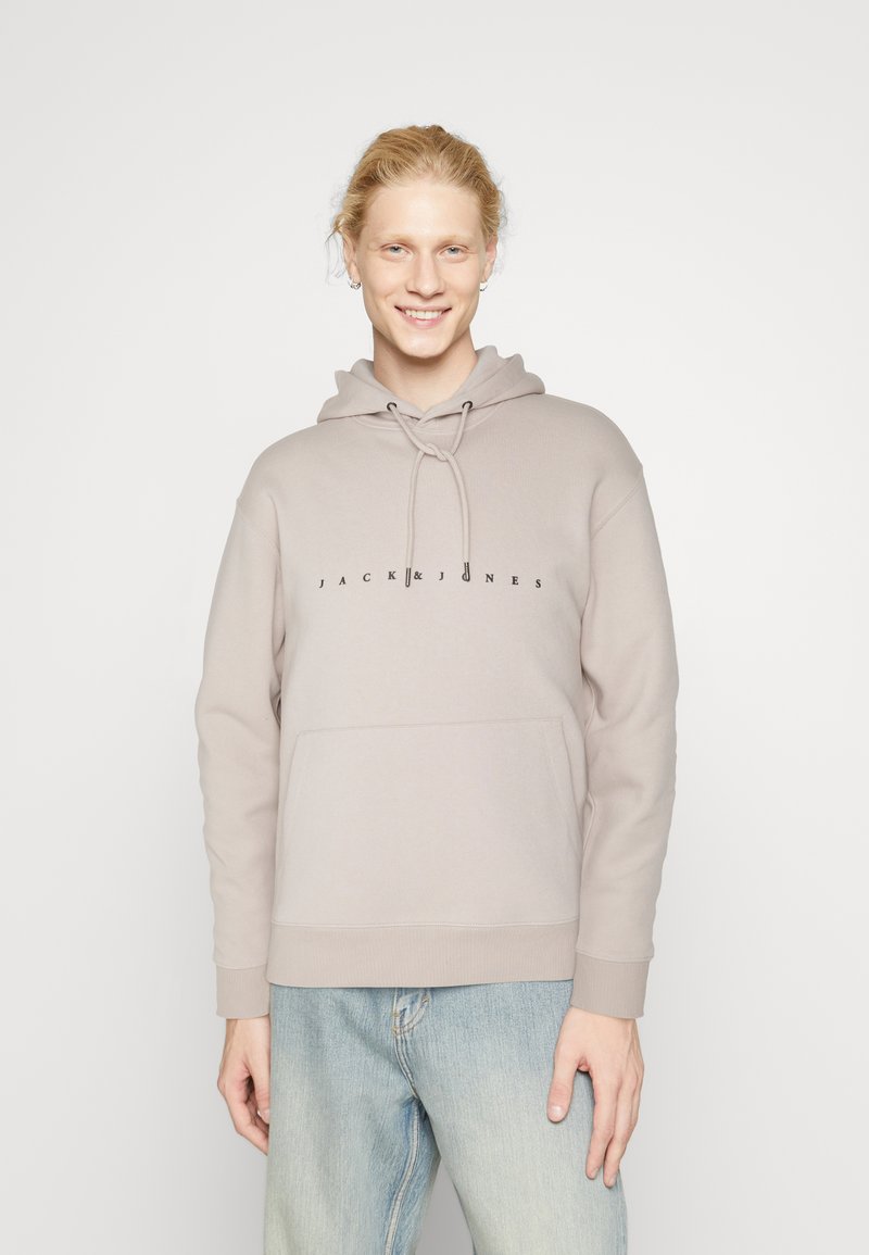 Šviesiai smėlinė vilnonių striukė su raišteliu, turinti priekinę kengūros kišenę ir juodą „JACK & JONES“ logotipą, įsiterpusį į krūtinę.