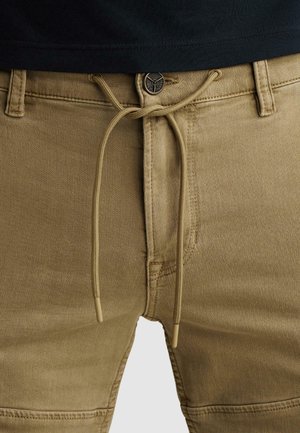 Primo piano di pantaloni beige con bottone, passanti per cintura e coulisse beige annodata in vita, indossati con una camicia nera.