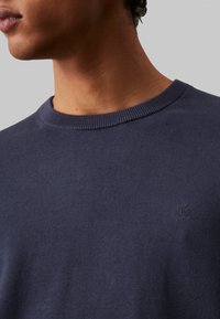 Maglione blu navy realizzato in tessuto a maglia, caratterizzato da un collo a girocollo e un piccolo logo impresso sul petto. Texture liscia con dettagli a coste.