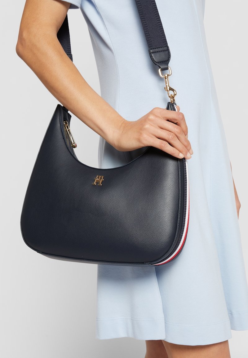 Femme portant une robe bleu clair tenant un sac à bandoulière en cuir noir avec des ferrures dorées et une bordure latérale rouge, blanche et bleue.