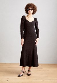 Sort langærmet kjole med tekstureret stof, tætsiddende bodice, subtil flair ved kanten og puffede skuldre. Båret med sorte spidse hæle med gulddetaljer.