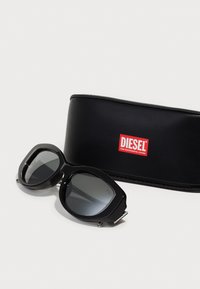 Black sunglasses with rounded frames, gray lenses, and silver accents; accompanied by a black case featuring a prominent red Diesel logo.