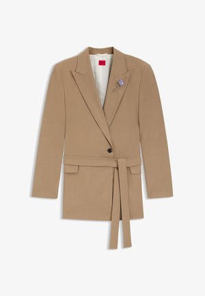 Blazer beige sur mesure avec ceinture, revers à encoches, fermeture à un bouton et épingle florale violette sur la poitrine. Texture de tissu lisse.