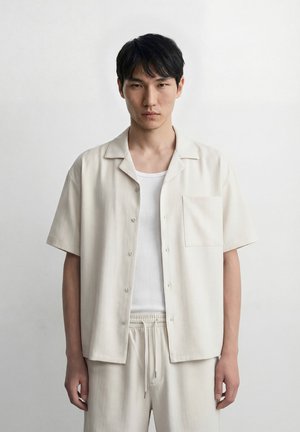 Junger Mann mit kurzen schwarzen Haaren, der ein weißes offenes Hemd über einem weißen Tanktop und passende helle Hosen trägt, vor einem einfarbigen Hintergrund steht.