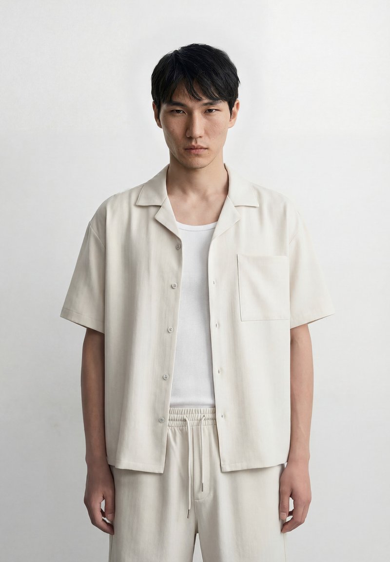 Jeune homme aux cheveux noirs courts portant une chemise blanche ouverte sur un débardeur blanc et un pantalon clair assorti, debout devant un fond uni.