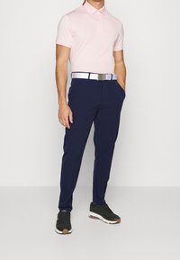 Ljusrosa polo skjorta med krage, marinblå byxor och ett vita bälte. Svarta sneakers med bruna detaljer, stående mot en enfärgad bakgrund.