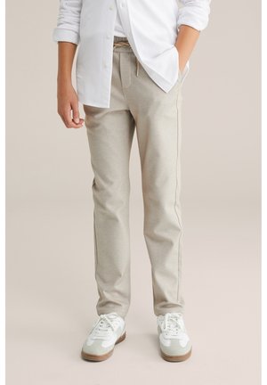 Person iført beige joggingbukser, hvide sneakers og en hvid langærmet skjorte med den ene hånd i lommen, stående på en neutral baggrund.