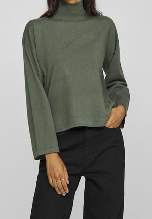 Personne portant un pull à col roulé vert olive et un pantalon noir, une main dans la poche et deux bagues aux doigts.