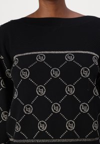 Pull en maille noire avec épaules côtelées, présentant un motif géométrique argenté et des accents de logo disposés en forme de losange sur le devant.