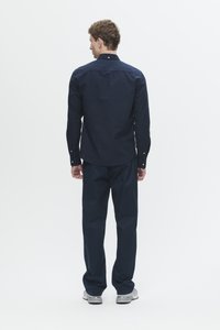 Man standing facing away wearing a navy long-sleeve shirt, navy pants, and white sneakers on a plain white background.