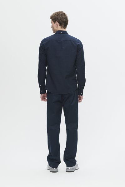 Homme de dos portant une chemise à manches longues marine, un pantalon marine et des baskets blanches sur un fond blanc uni.