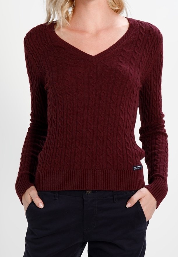 Femme portant un pull en maille torsadée bordeaux à col en V avec manches longues et un pantalon noir, mains dans les poches.