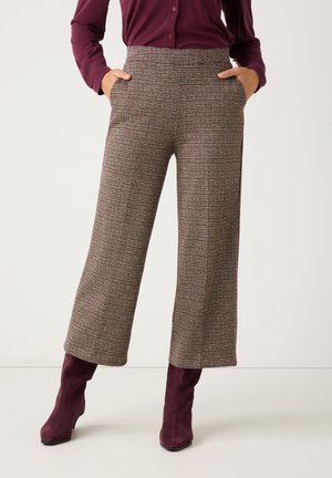 Kobieta nosząca wysokie, szerokie spodnie w kratę z rękami w kieszeniach, w połączeniu z bordową koszulą z długim rękawem i dopasowanymi butami na obcasie.