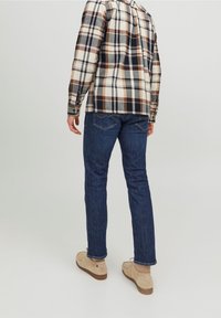 Camicia in flanella a quadri nei colori beige, blu navy e marrone; abbinata a jeans a gamba dritta blu scuro e scarpe beige, vista da dietro.